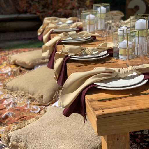 rustic-pallet-picnic-table-glass-candle-boxes-with-champagne-and-burgundy-linen-napkins-AFC-crockery-and-standard-glassware-assorted-cushions-and-wandering-folk-rugs