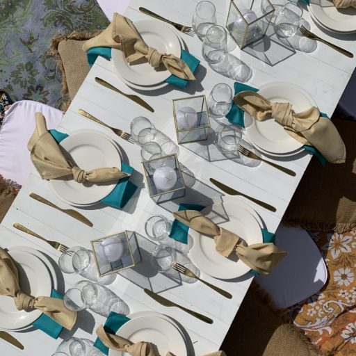 overhead-view-of-white-pallet-picnic-table-glass-candle-boxes-with-champagne-and-turquoise-linen-napkins-AFC-crockery-and-standard-glassware-assorted-cushions-and-wandering-folk-rugs